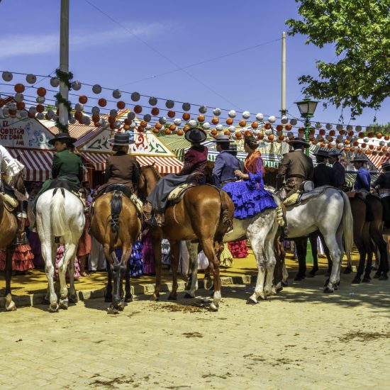 Seville, Andalusia, Spain, Europe - April 14, 2024. Seville April Fair fairgrounds with horseback riders, women in suits and traditional Spanish flamenco dresses on horseback, some riding side saddle, and men in suits and hats. The festival goers socialize, drink and eat on horseback in front of colorful striped entertainment party tents on a decorated city street lined with festive banners. Thousands of tents are constructed for the annual Spanish cultural heritage festival, the April Fair, and the celebration lasts for seven days. The fair is also an equestrian event with a daily parade of decorated horse-drawn carriages, horseback riders as well as entertainment, music, food, drink and community.